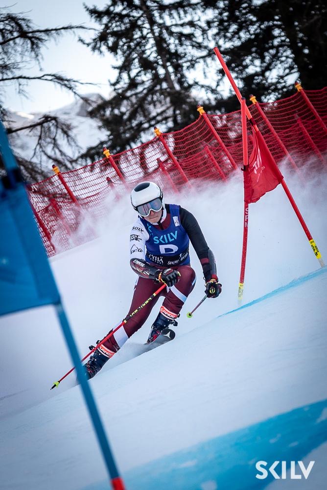 Alpine skier in a professional maroon and white racing suit navigating a slalom course through red gates during a competition.