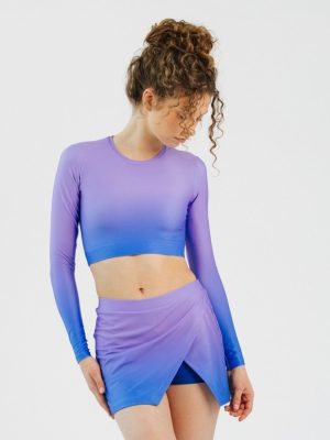 A model showcases a custom-designed overlapping cheerleader's skort and matching long-sleeve crop top, featuring a vibrant purple-to-blue ombre gradient on technical performance fabric.