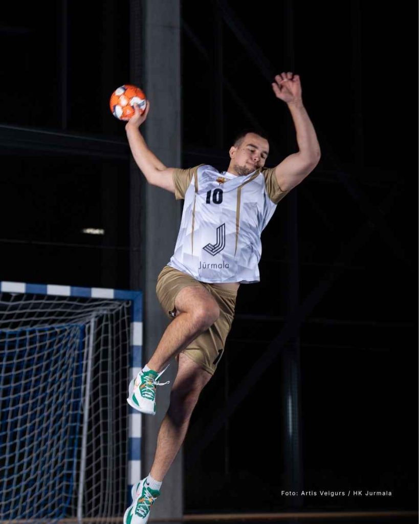 Handball player wearing custom white and gold HK Jūrmala jersey mid-air jumping to throw a handball.