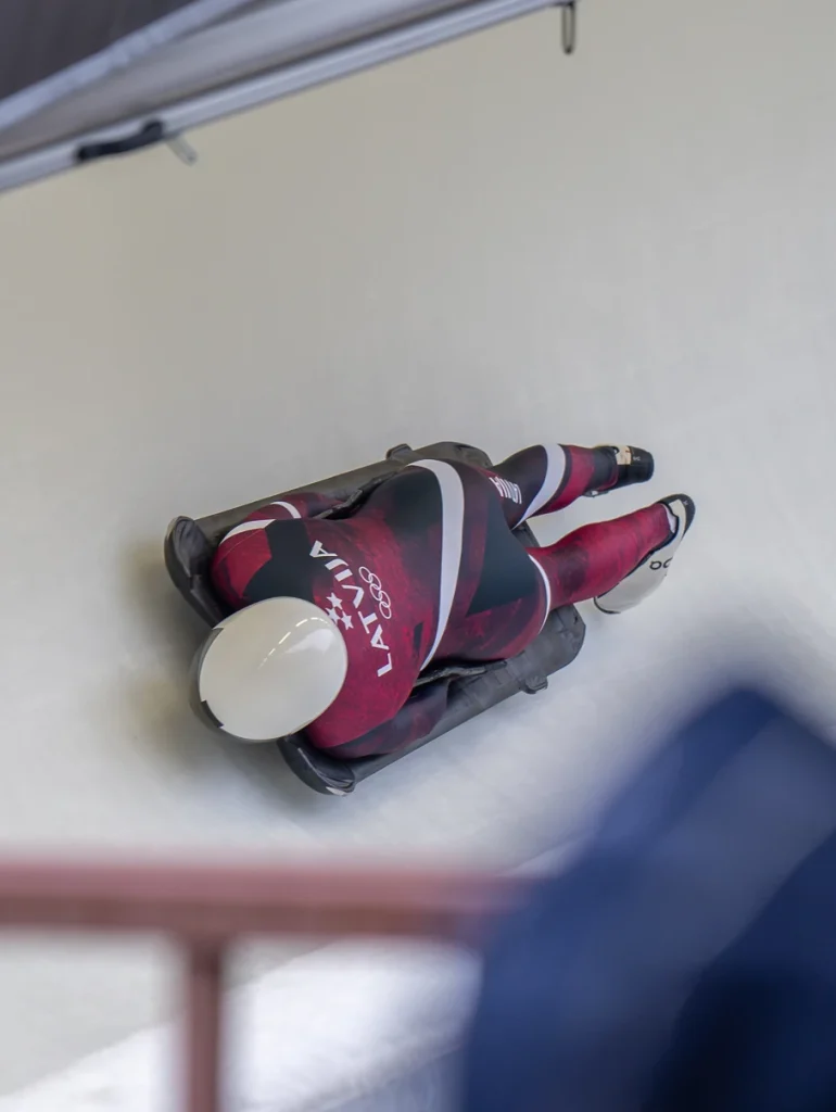 An action shot of an elite athlete in a custom teamwear skeleton speed suit navigating an ice track, showcasing the aerodynamic performance and high-definition sublimation printed "Latvija" branding and Olympic rings on specialized technical fabric.