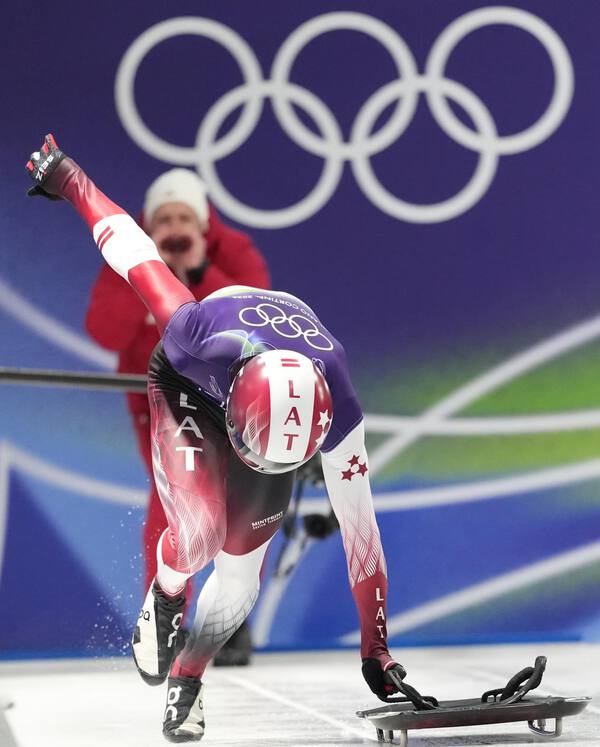 An action shot of a Latvian skeleton athlete at the Winter Olympics, wearing a high-performance custom teamwear speedsuit. The aerodynamic suit features professional sublimation printing with national symbols and "LAT" branding, designed for maximum speed and precision on the ice.