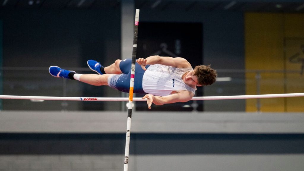 A male athlete in action wearing a custom-designed, sublimated pole vault kit. The high-performance athletic wear features personalized graphics and a professional fit, shown during a high-clearance jump in a stadium.