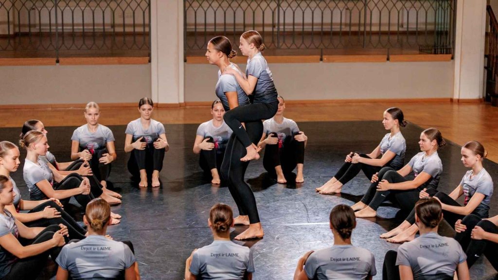 Dancers from dance studio Dzirnas train in studio apparel, wearing grey gradient performance shirts with custom "Dejot ir LAIME" branding.