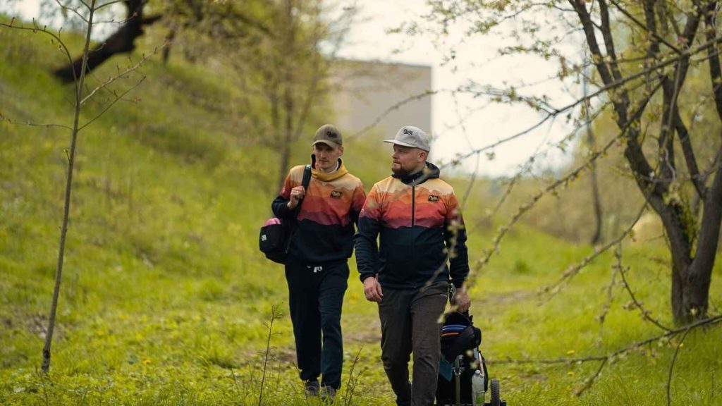 Two disc golf players walk through a wooded course wearing matching Chilli Disc Golf custom team hoodies