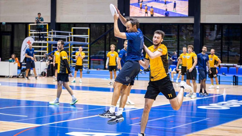Professional action shot from an indoor ultimate frisbee match, featuring custom sportswear team jerseys with blue and yellow sublimation print designs.