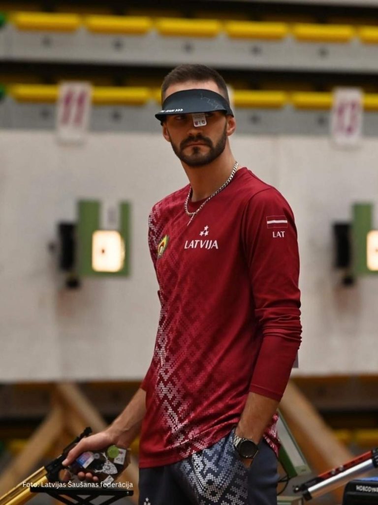 Professional marksman from the Latvian national shooting team wearing a custom maroon jersey with "LATVIJA" branding and national symbols during a competition.