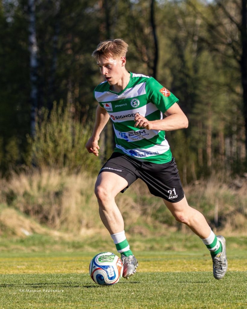 Action shot of a Maskun Palloseura football player in a green and white custom jersey dribbling a soccer ball on a grass pitch.