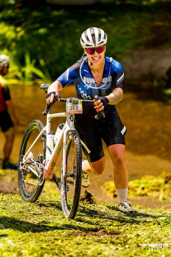 Female cyclist in custom blue Junda Racing MTB cycling jersey pushing gravel bike.